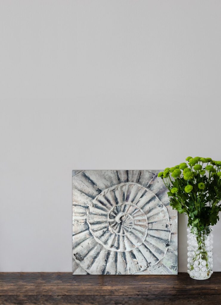 The painting of a shell standing on wooden floor, next to a vase of green flowers.