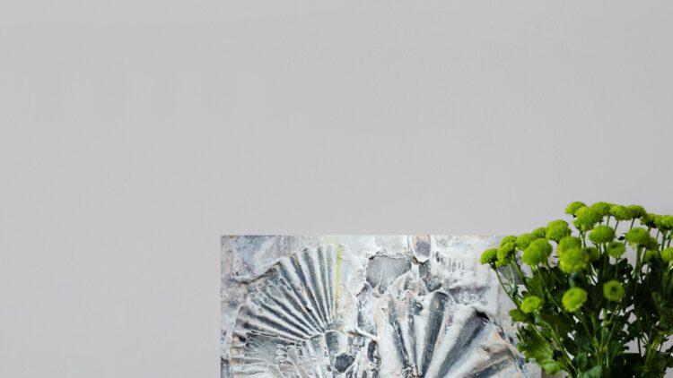 The grey shell painting standing on the wooden floor, next to a flower vase.